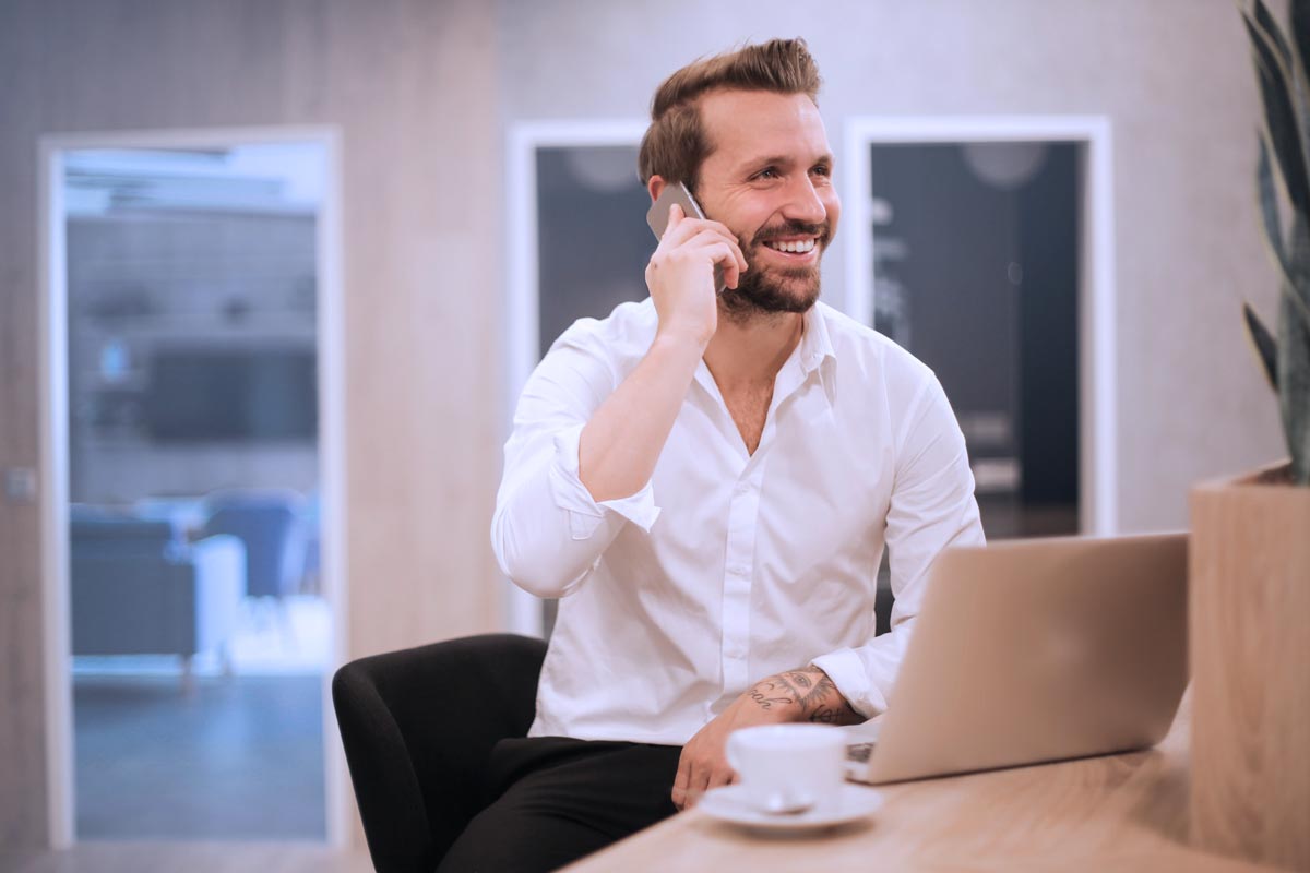 a man sits smiling in front of his laptop and talking on the phone