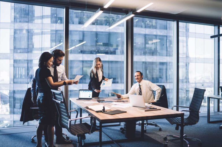 two women and two men sit and stand around a conference table, looking at documents and laptops