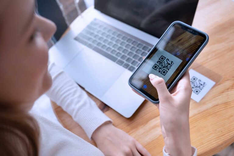 A woman is sitting at an office desk with a laptop on it. The woman is holding a smartphone in her hand and uses it to scan a QR code lying on the table in front of her.